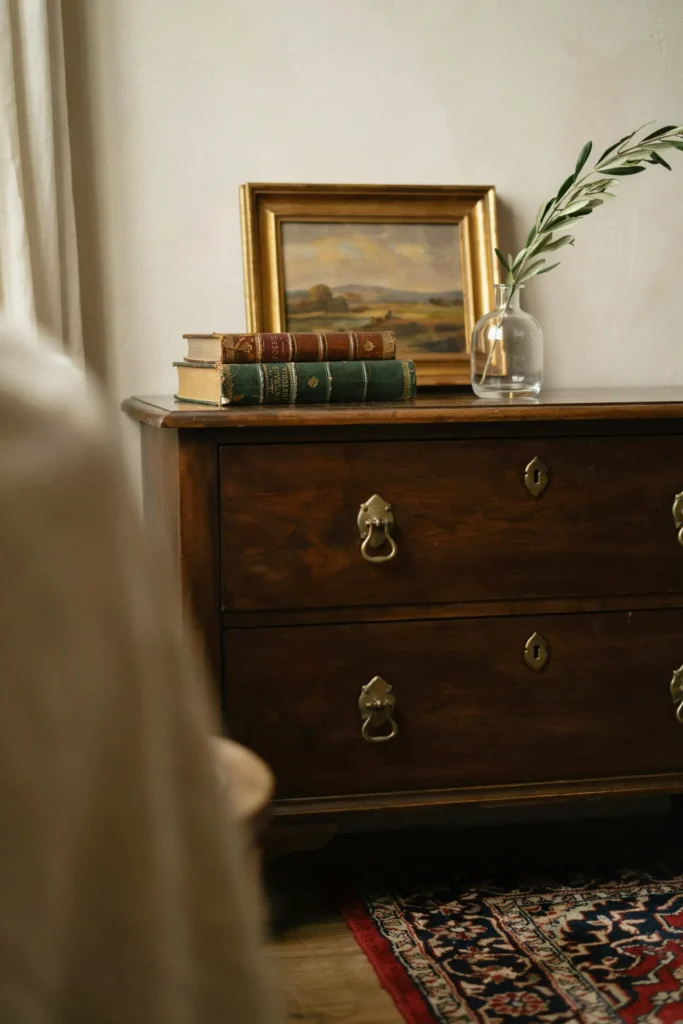 A styled close-up shot of an eclectic, transitional bedroom corner featuring a rich history. A vintage, dark walnut wood chest of drawers with aged brass drop handles stands beautifully against a warm cream wall. The color palette blends deep wood tones, rich golds, and soft ivory. Atop the chest rests a stack of leather-bound books, a small oil painting in a gilded frame, and a clear glass vase holding a single green olive branch. The atmosphere is soulful, curated, premium, and full of character.