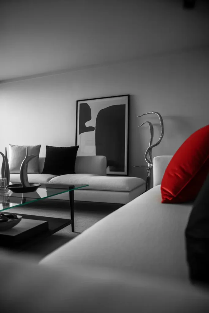  A sleek modern living room in monochrome style, soft diffused lighting with subtle natural highlights, featuring black, white, and grey tones with bold red accents. Minimal sofa, glass table, abstract art, and metal decor. Smooth textures with sharp contrasts. Clean sophisticated mood. Wide shot with strong foreground styling and crisp background layering.