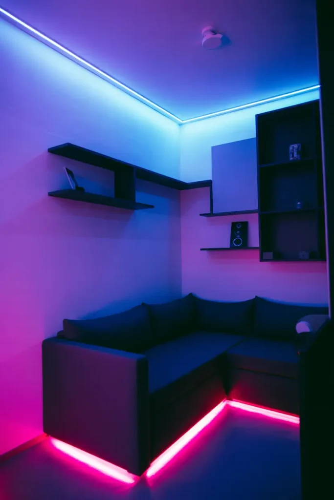 Modern college apartment living room corner with LED strip lighting, neon accents, low ambient lighting with purple and blue tones, sleek minimal furniture, black and grey palette with vibrant light contrast, small sofa, wall-mounted shelves, tech-inspired decor, glowing edges behind furniture, cozy night vibe, angled corner shot highlighting lighting depth and layered glow effect.